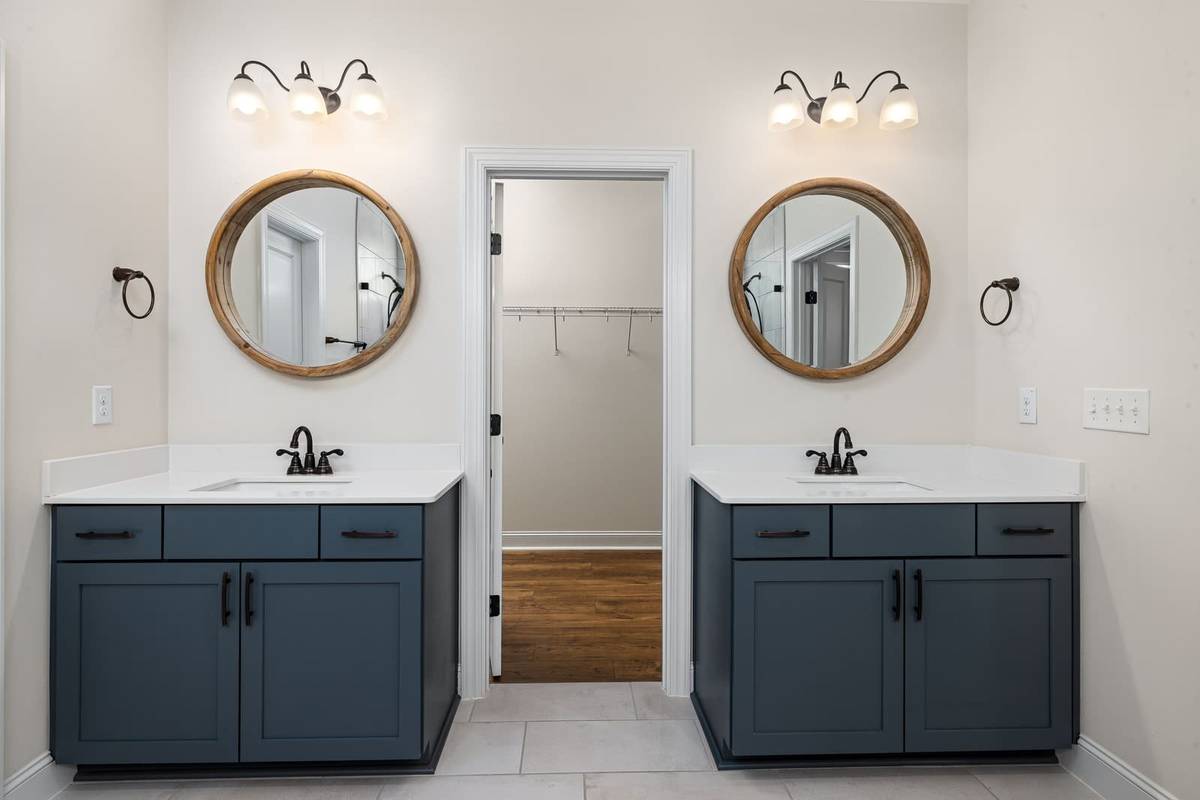 Two slate blue vanities with white tops, each under a round wood-framed mirror and a three-light fixture