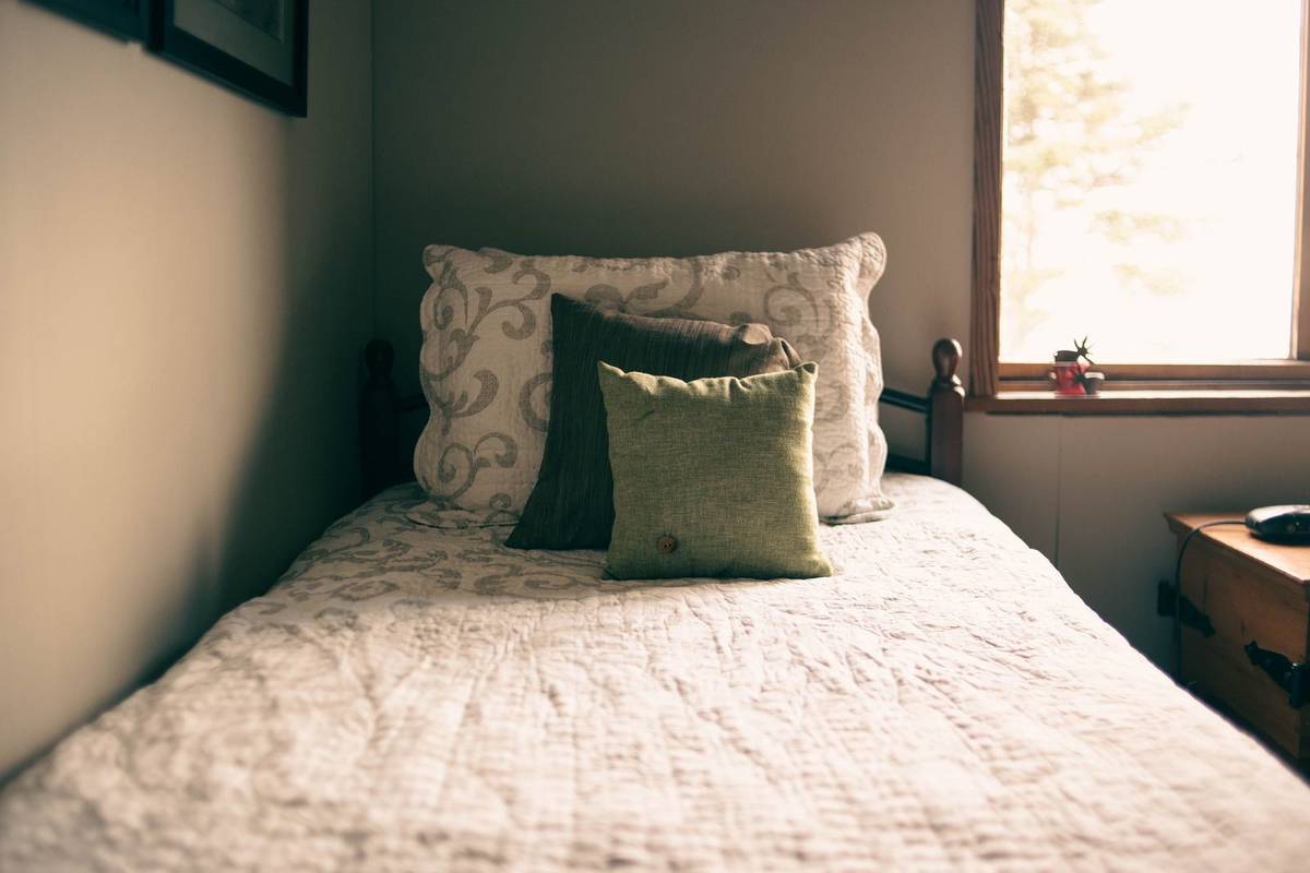 A twin bed made with a white quilted coverlet, a matching sham and two small accent pillows by a window