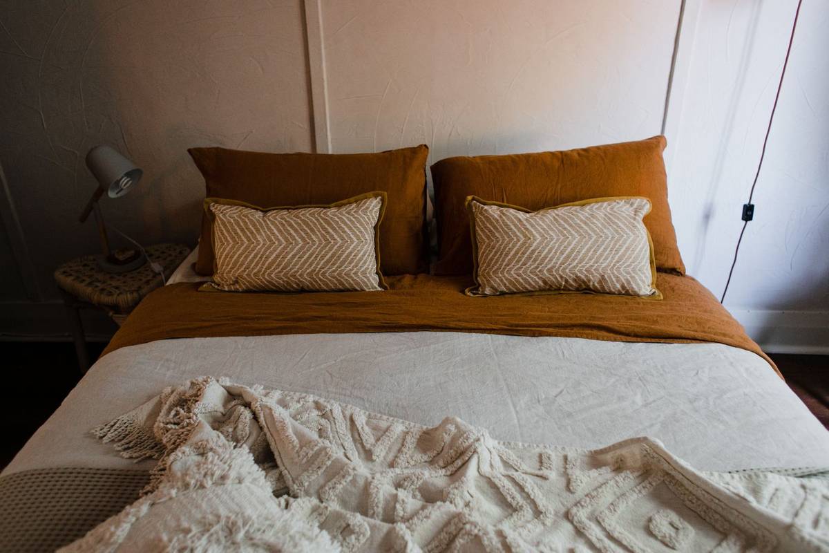 Rust linen sheets and pillowcases under a cream coverlet and a tufted throw, with two patterned pillows