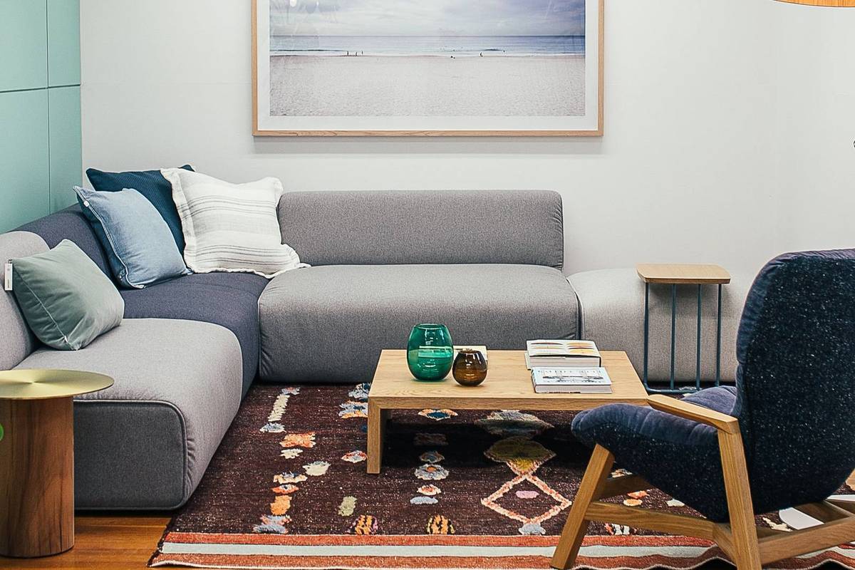 A gray sectional sofa behind a light wood coffee table on a patterned rug, with a framed beach print above