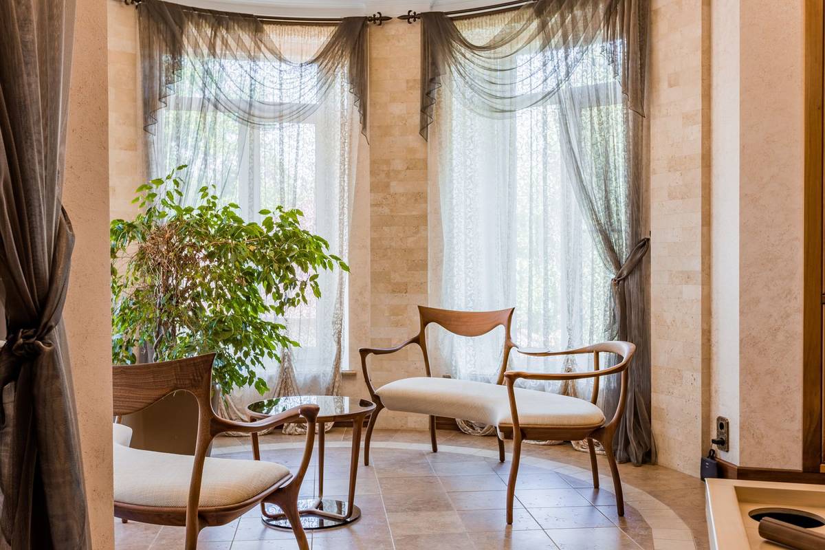 A bay window with sheer panels and draped valances on each window, over two chairs and a small round table