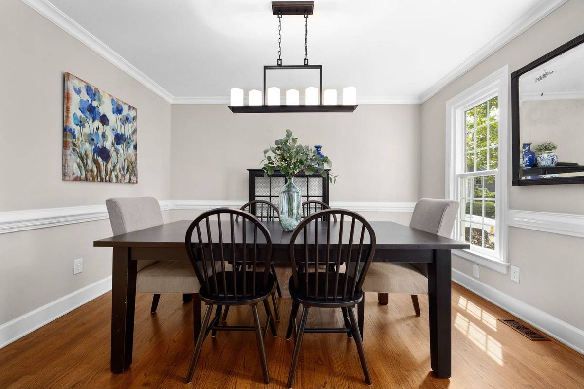 A long linear light with candle-style bulbs over a dark wood table and six chairs, with a floral painting on the wall