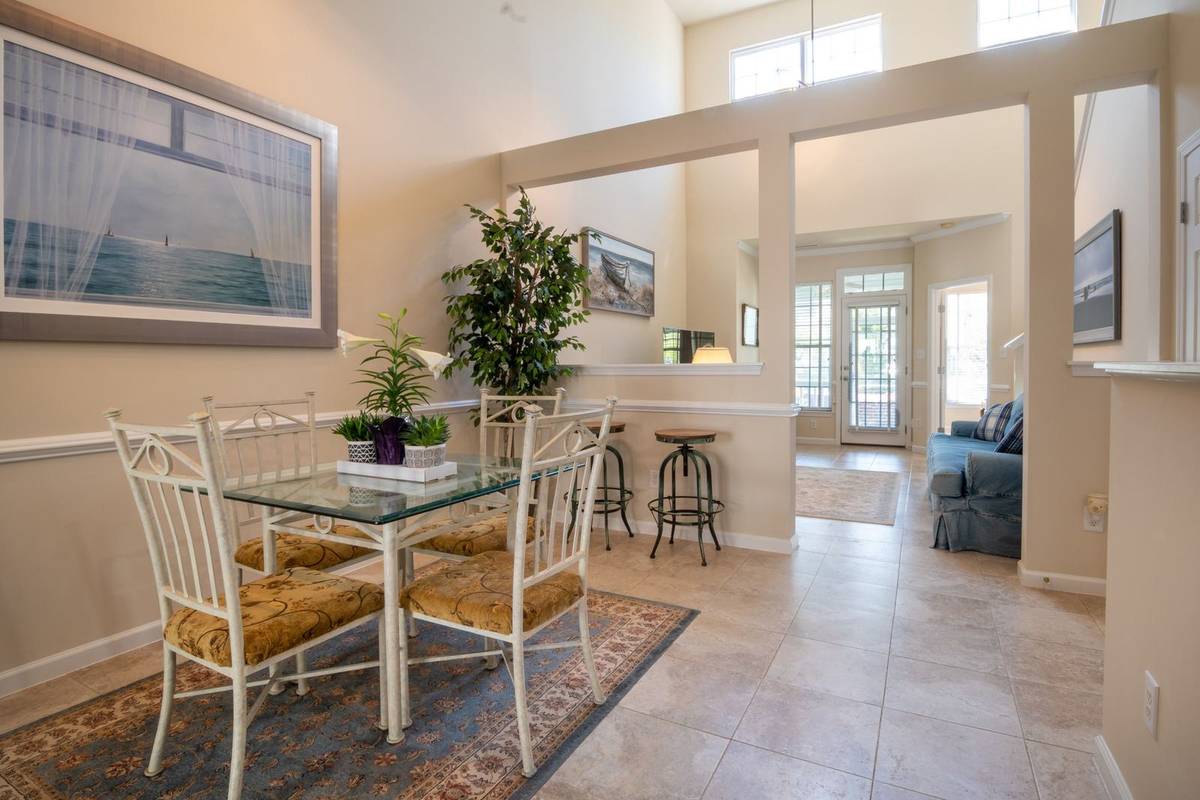 A glass table and four chairs on a patterned rug at one end of an open room, with the living area beyond