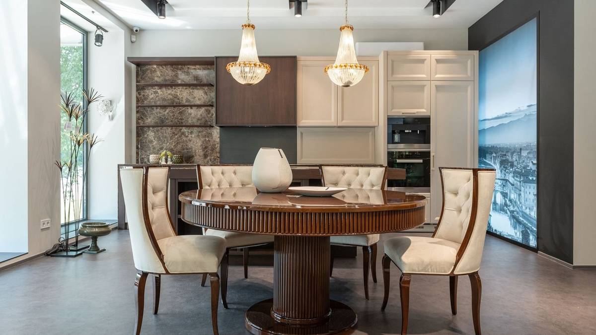 A round pedestal dining table with upholstered chairs under two crystal chandeliers