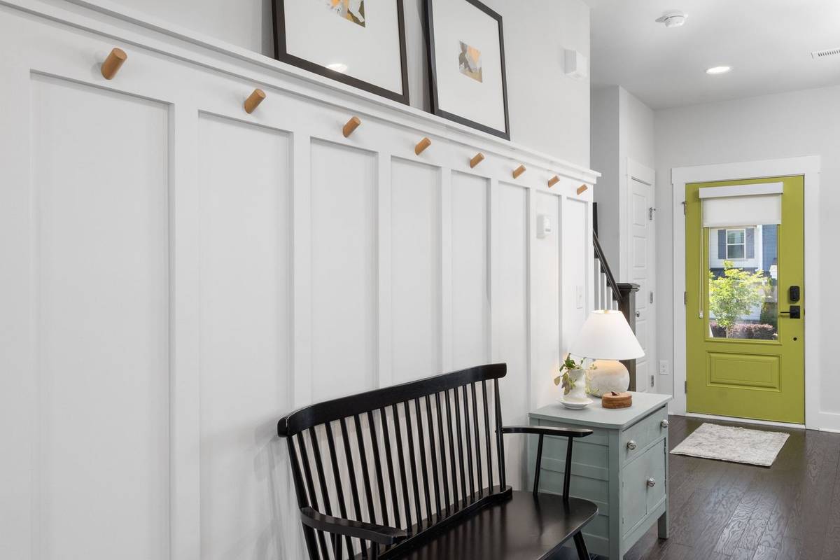 A black spindle bench under a row of wood peg hooks on a paneled wall, a small rug at a green front door beyond