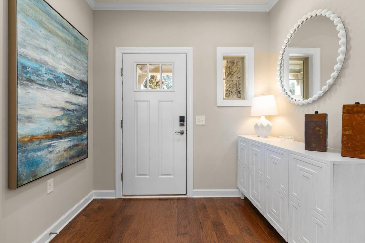 A long white console with a lamp and two boxes beside the front door, a round mirror above it and a large painting across