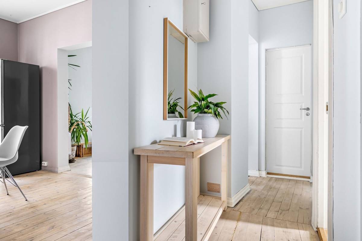 A slim wood console with a plant and an open book under a narrow mirror, on a short wall in a light hallway