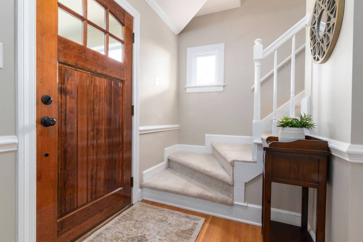 A wood front door with a small rug before it, a narrow wood console table with a plant, and carpeted stairs beyond