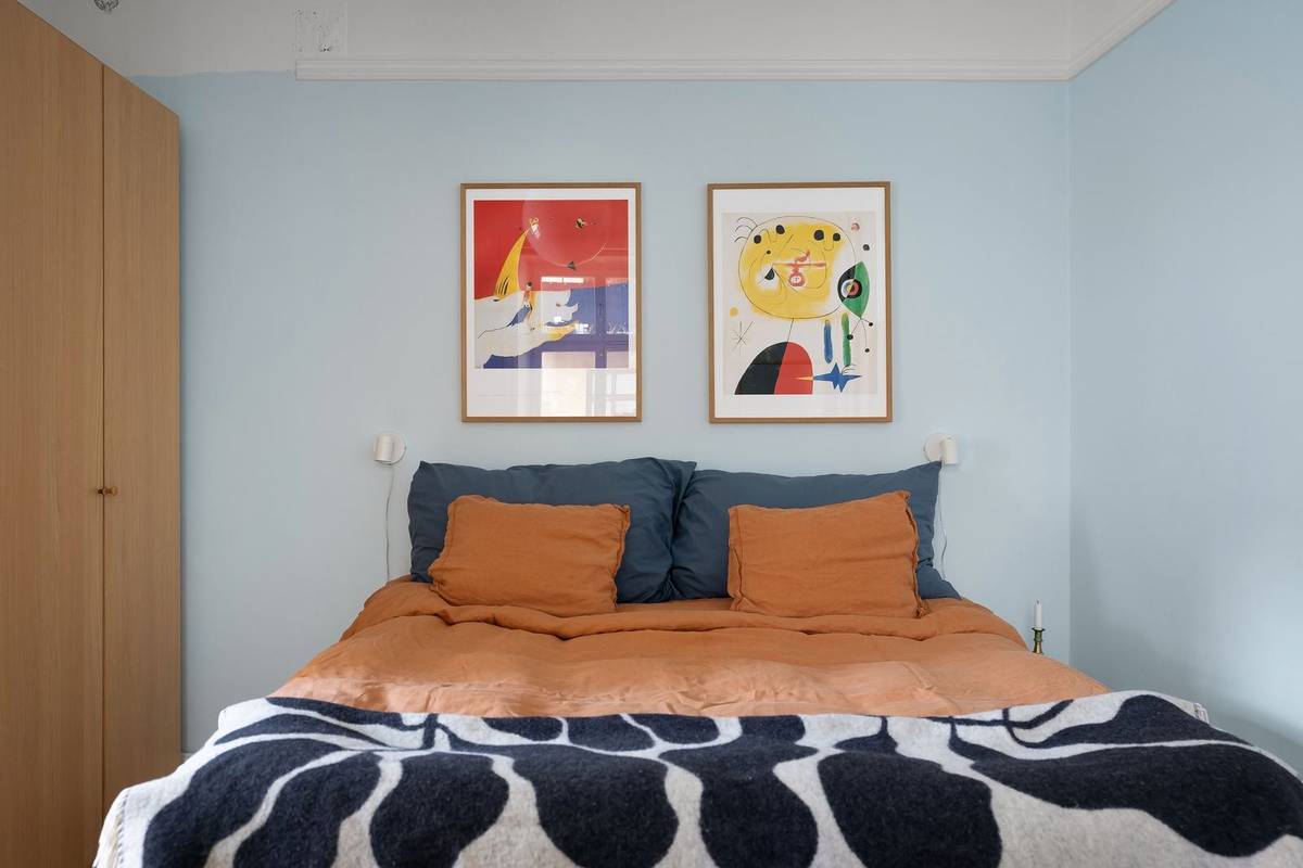 A bed with no headboard against a pale blue wall, two framed abstract prints above it and a wall lamp at each side
