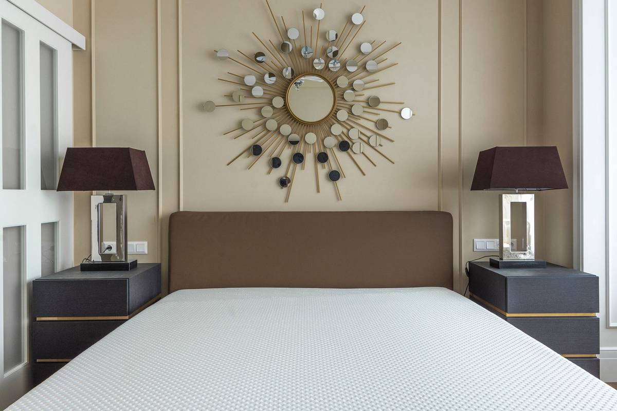A sunburst mirror centered above a low brown upholstered headboard, between two dark nightstands with table lamps