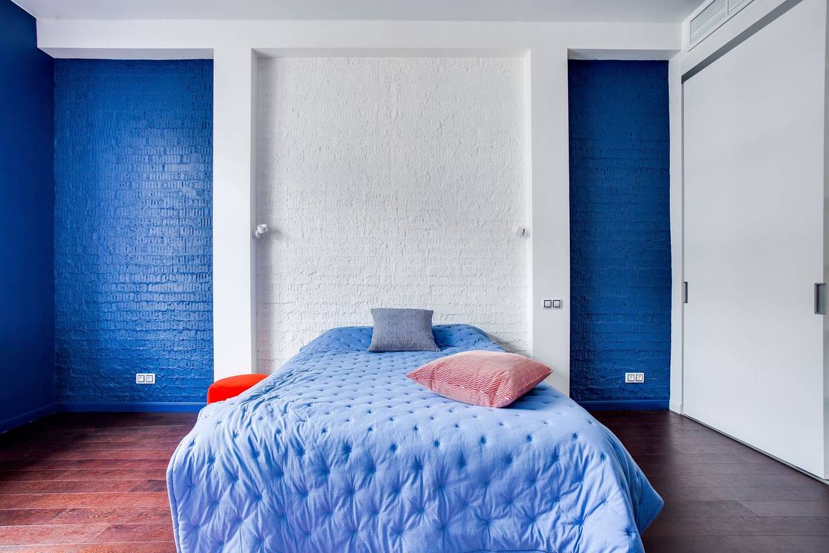 A bed with no headboard in front of a white painted brick panel framed by deep blue walls, like a headboard