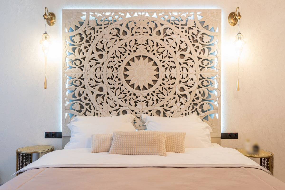 A carved white wood panel hung on the wall behind the bed as its headboard, lit from behind, with sconces at each side