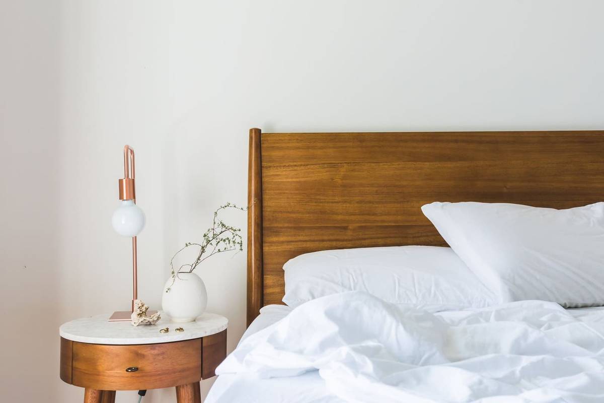 A bed with a walnut wood headboard and white bedding, beside a round wood nightstand with a copper table lamp