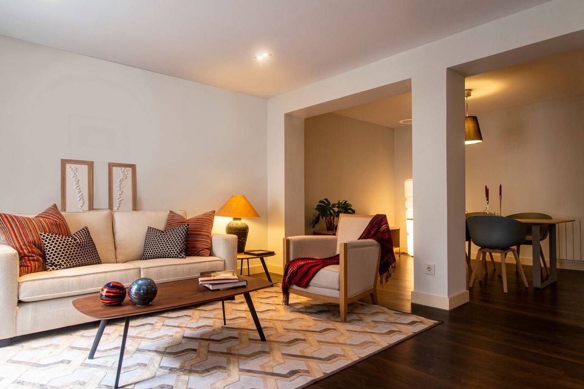 A living room corner with a cream sofa, patterned pillows, a lit table lamp and a geometric rug on a wood floor