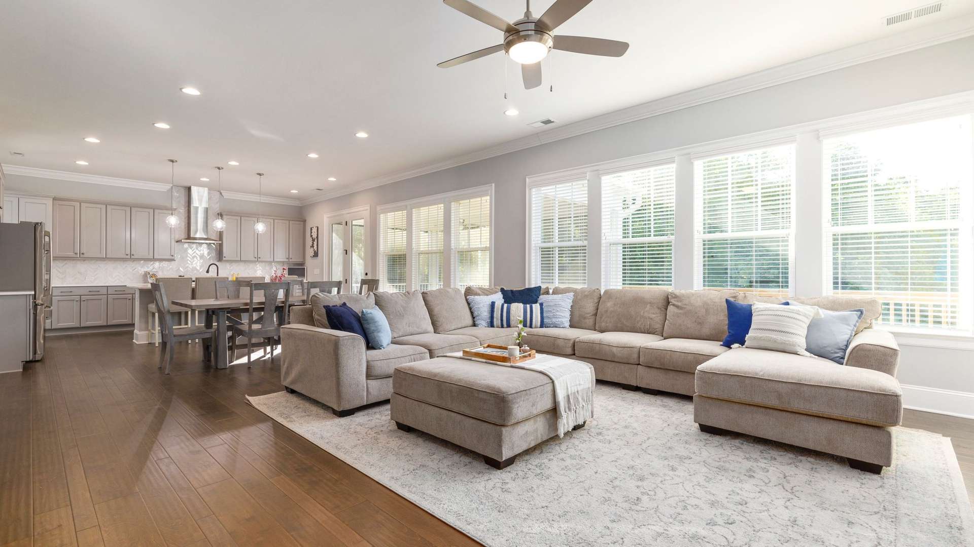 An open living room with a gray sectional, blue pillows and a pale rug, the kitchen and a wall of windows beyond