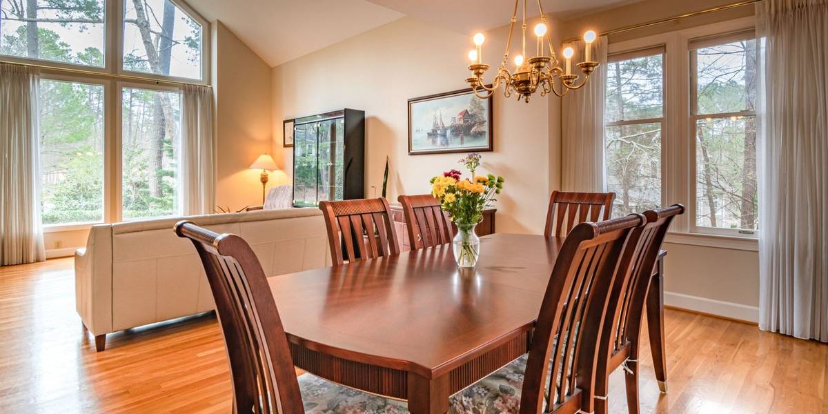 A dining room with a wood table, tall chairs and a brass chandelier, with a sofa and tall windows beside it
