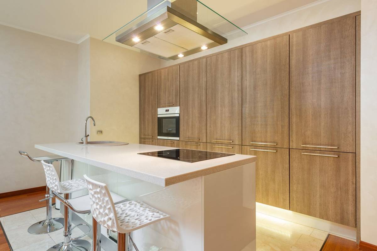 An island with a cooktop and a sink, a glass range hood above it and white stools along the far side