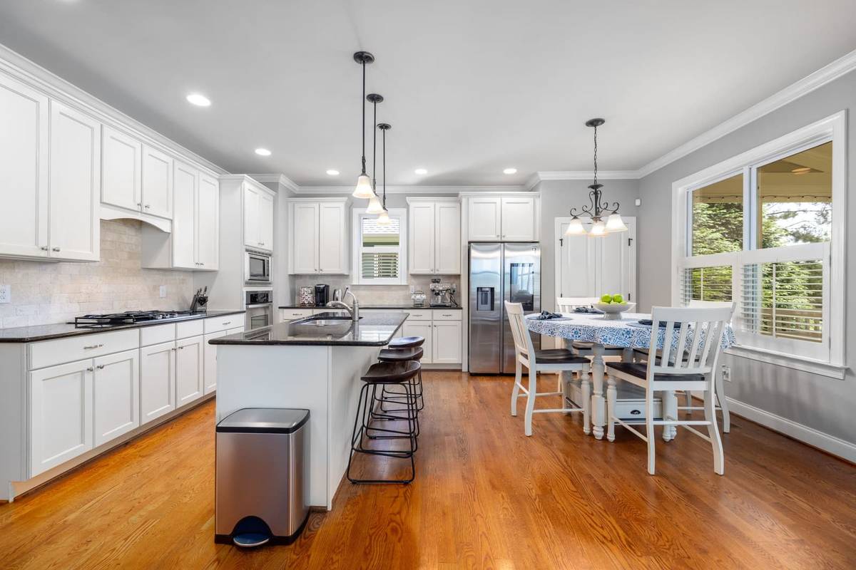A white kitchen with a long dark granite island and three counter stools along one side, with open floor around it