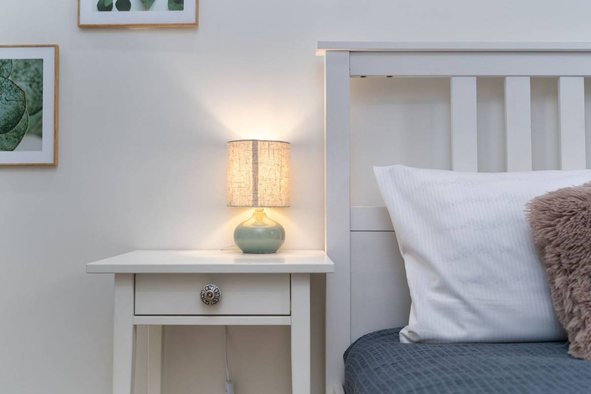 A white nightstand with one drawer and a small lit lamp beside a white bed with pillows, its top a little above the mattress