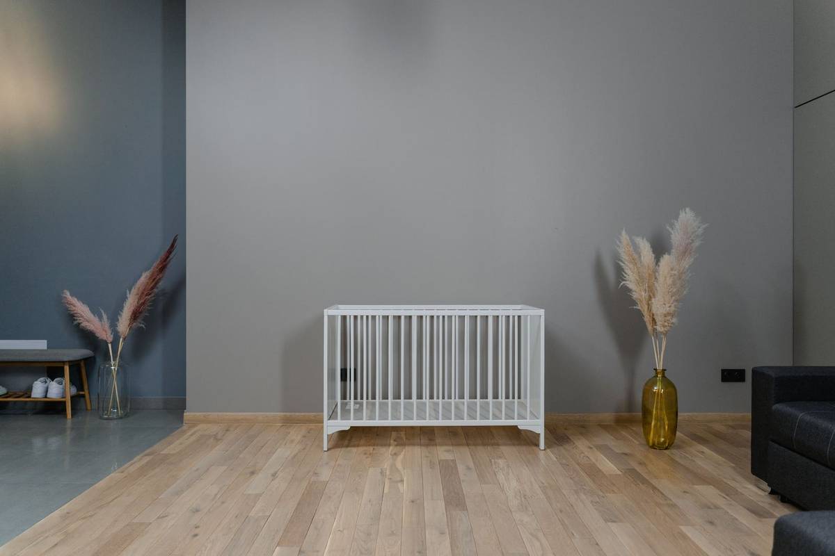 A plain white crib with nothing in it against a gray wall, with dried pampas grass in vases on the floor