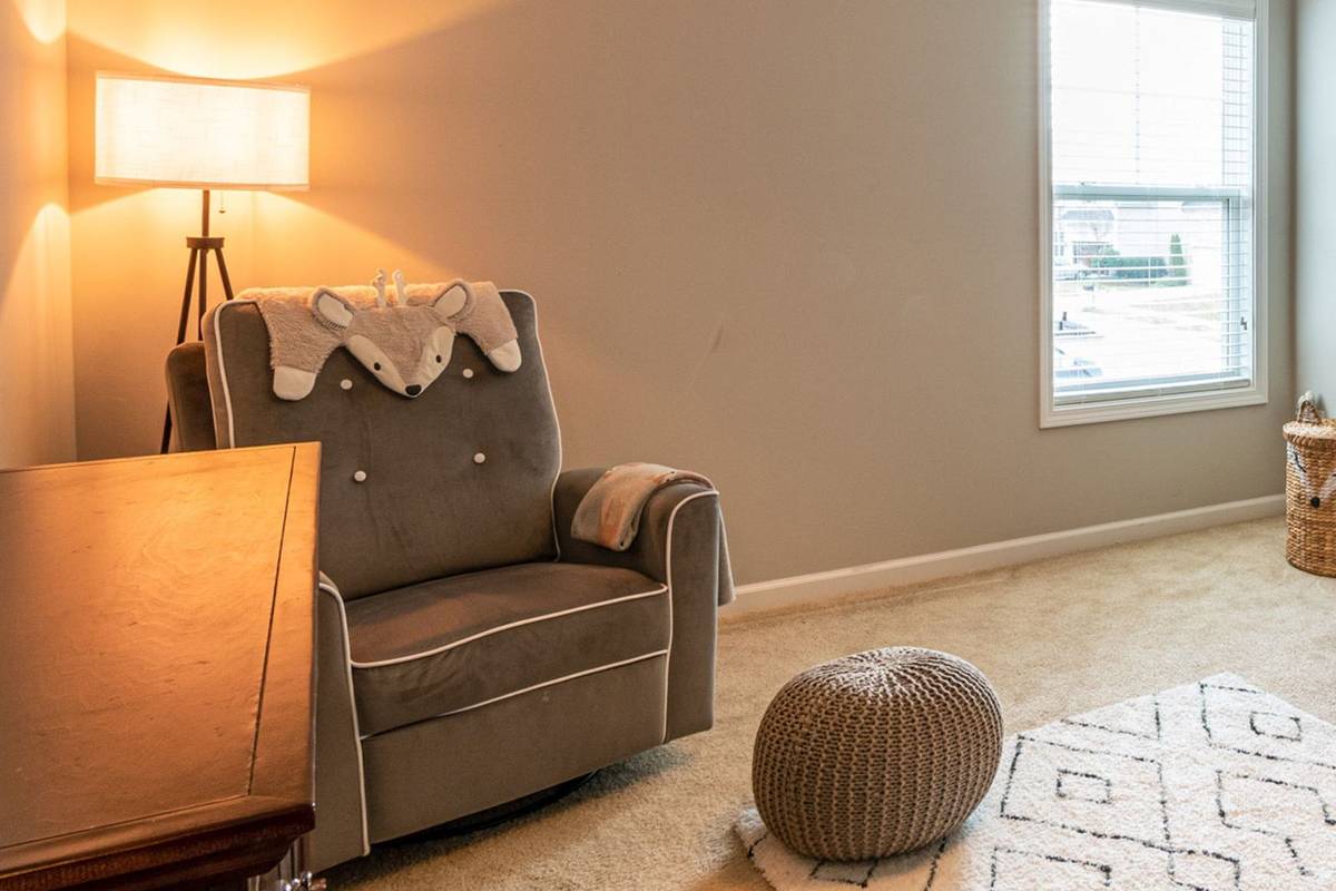 A gray glider in the corner of a nursery with a floor lamp behind it, a knit pouf and a patterned rug