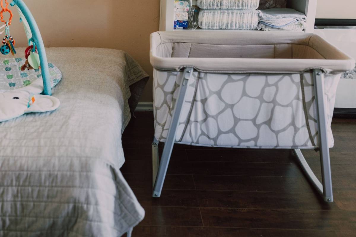 A bassinet with bare mesh sides beside an adult bed, with diapers and blankets on the changing table behind it