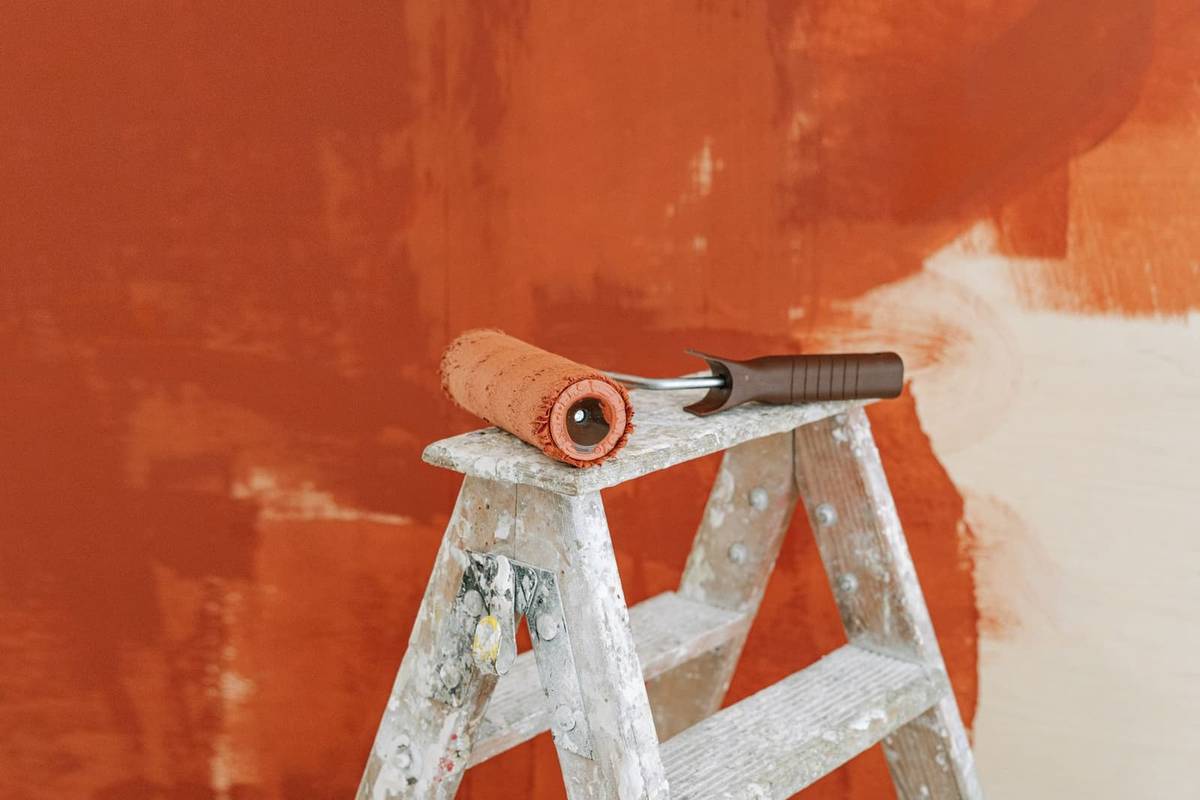A paint roller resting on a paint-spattered wooden stepladder in front of a wall half painted in terracotta orange