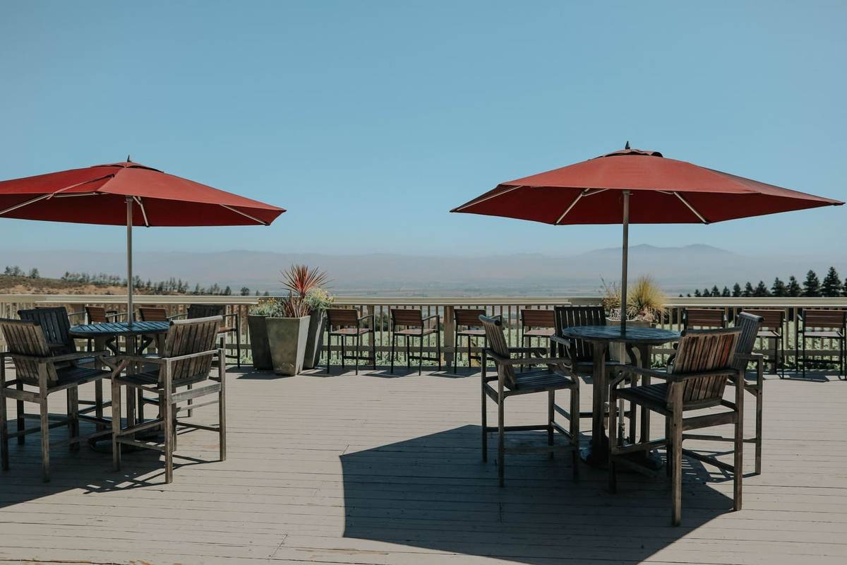 Two round tables with wooden chairs on a deck, each under a red market umbrella reaching past the table's edge