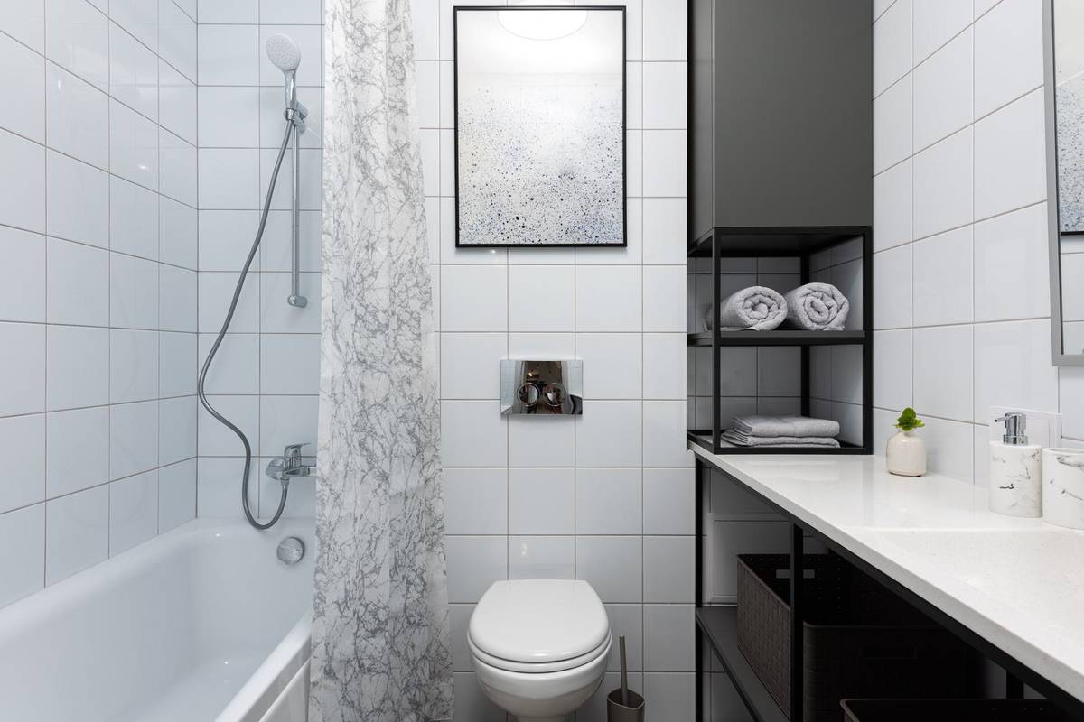 A narrow bathroom with a tub and shower curtain, a wall-hung toilet and open shelves of towels above a long counter