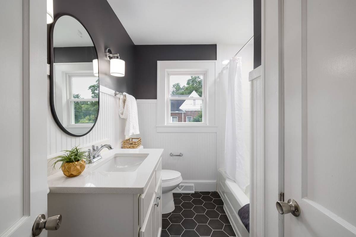 A narrow white vanity along one wall of a small bathroom, an oval mirror and a sconce above it, the toilet and a tub beyond