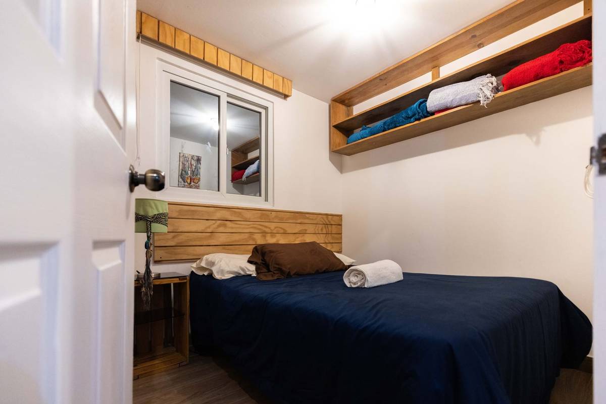 A small bedroom with the bed pushed into a corner under a wood headboard and open shelves of blankets high on the wall