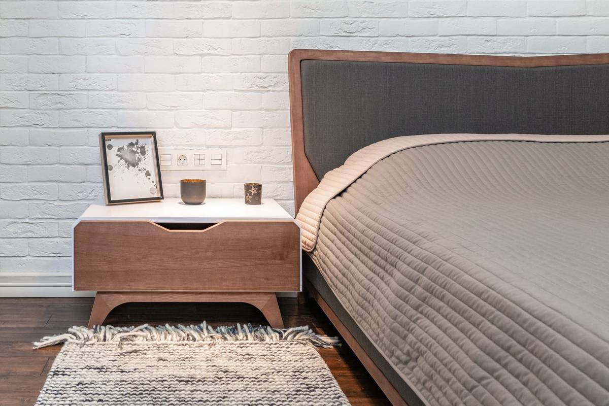 A small wood nightstand with a framed print and two candles, its top just above the mattress, and a woven rug in front
