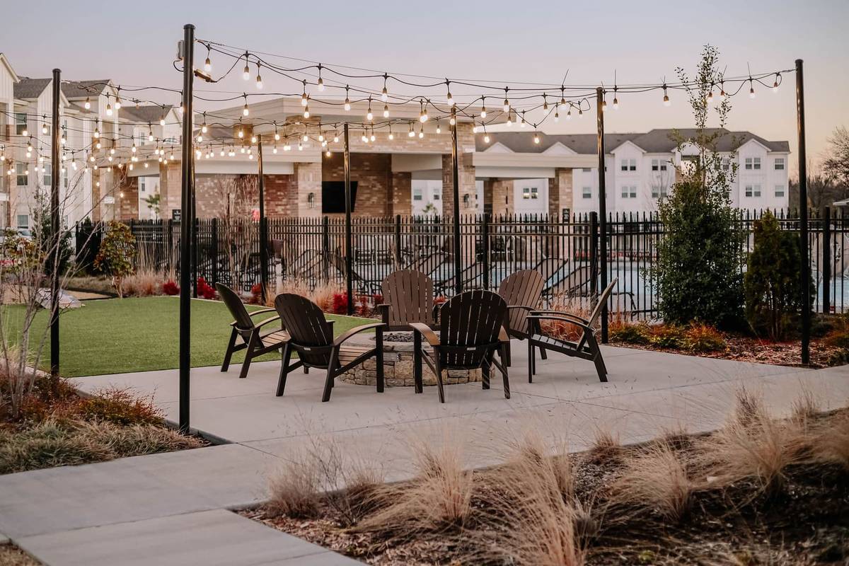 Patio string lights hung between black posts at dusk in zigzag runs over a fire pit ringed by wooden Adirondack chairs