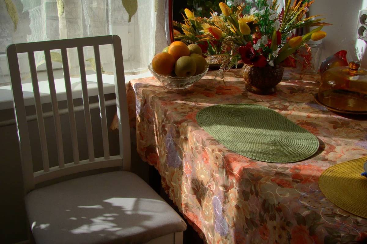 A table by a sunny window under a floral tablecloth that hangs over its edge, with place mats and a bowl of fruit