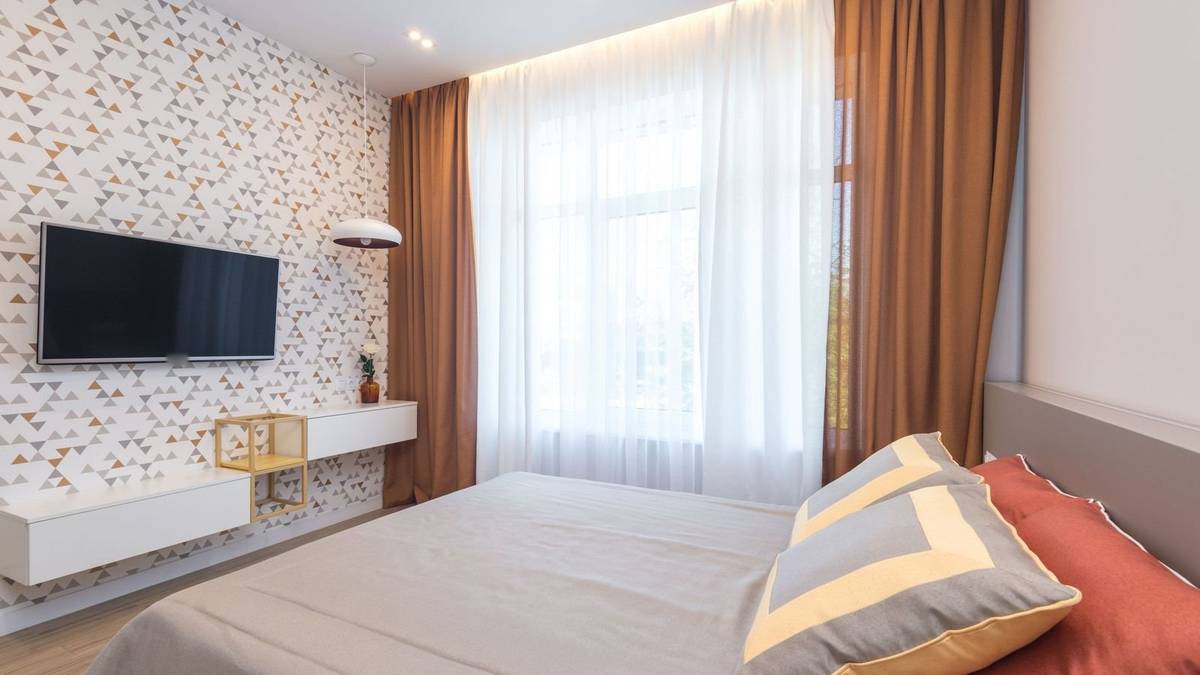 A TV on a patterned bedroom wall above a white floating shelf, with the bed in front