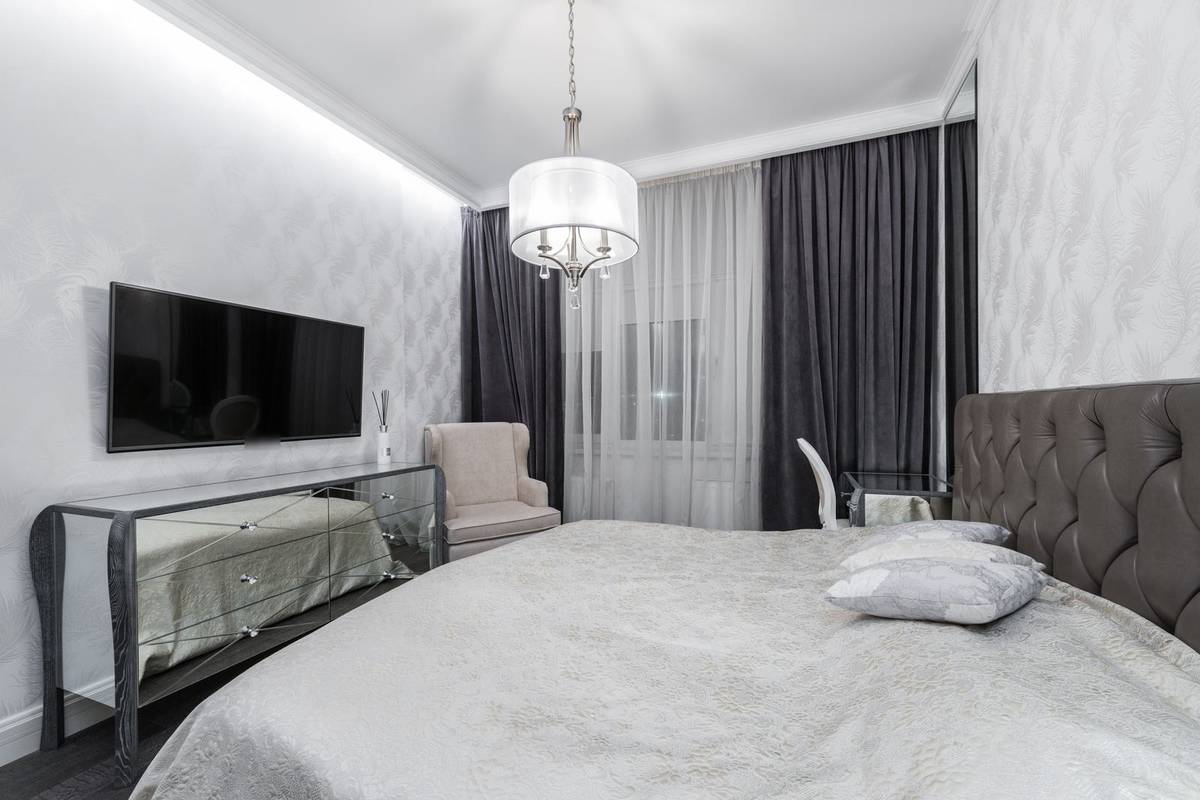 A bedroom with a TV mounted on the wall above a mirrored dresser, across from an upholstered bed