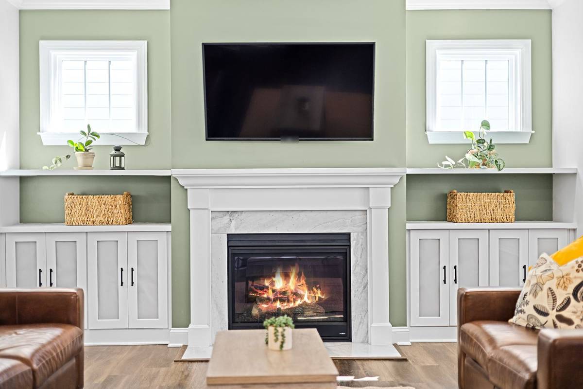 A TV mounted above a white fireplace mantel on a sage green wall, between built-in cabinets and two windows