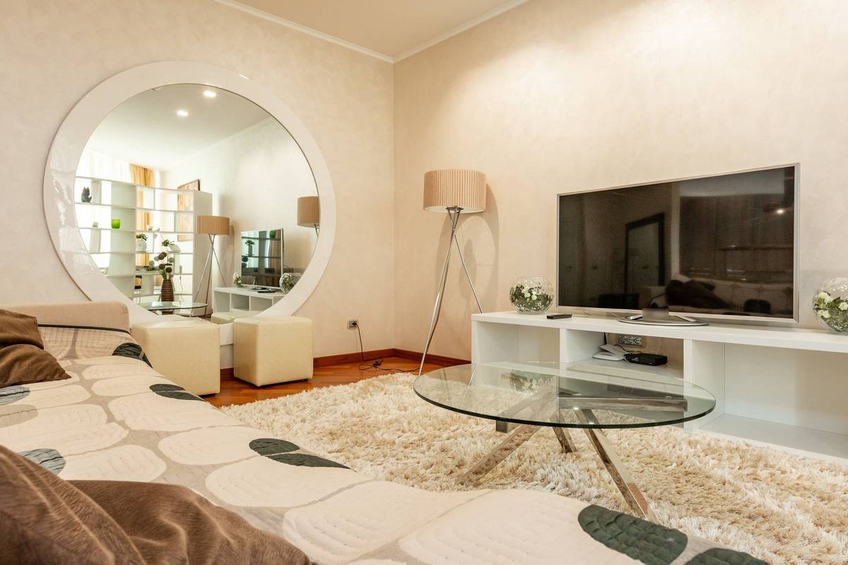 A TV on a pedestal base on a long white stand much wider than the screen, beside a tall floor lamp